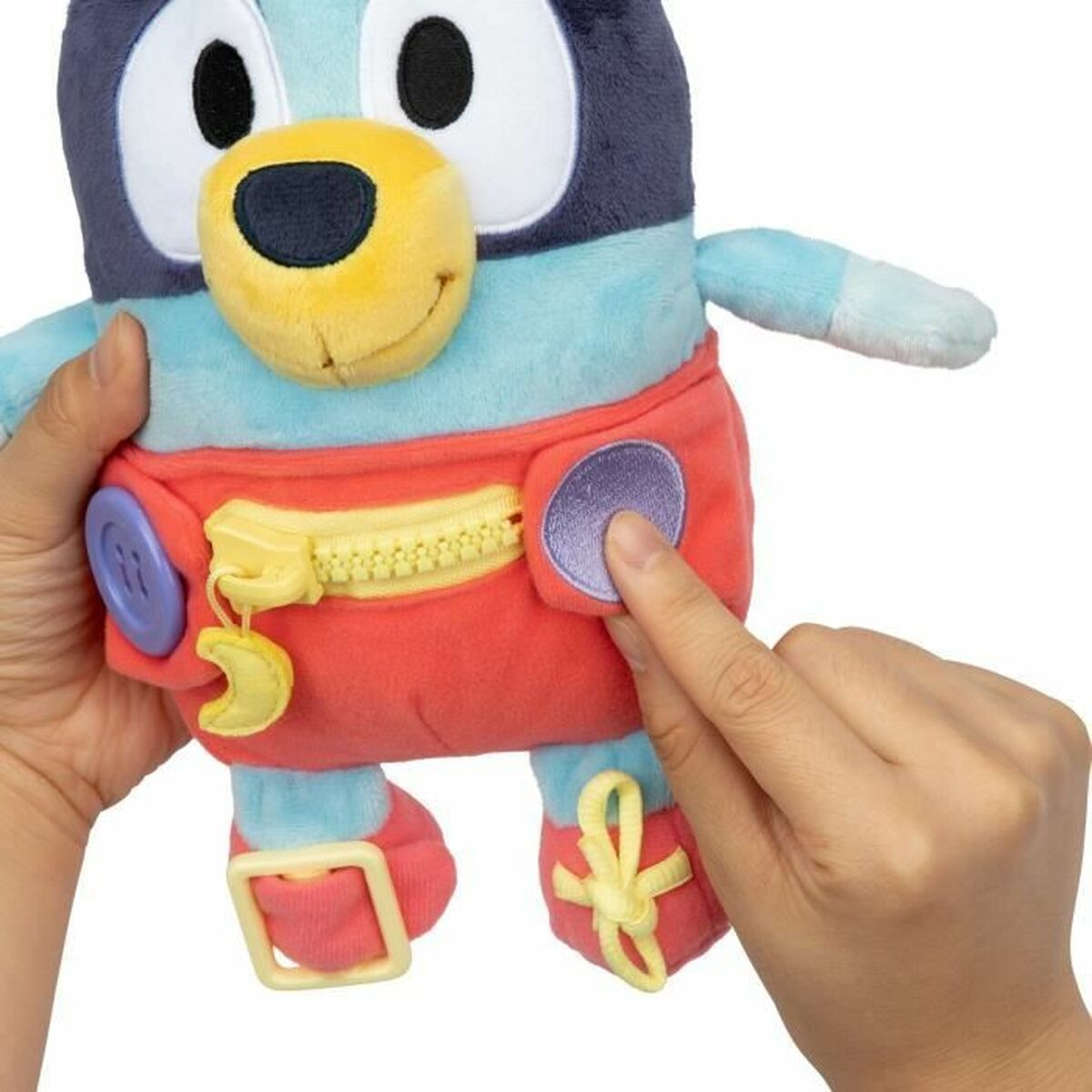 Une peluche Bluey est tenue par des mains. Le jouet a un pantalon rouge avec une fermeture éclair jaune, et un bouton violet est en train d'être pressé.