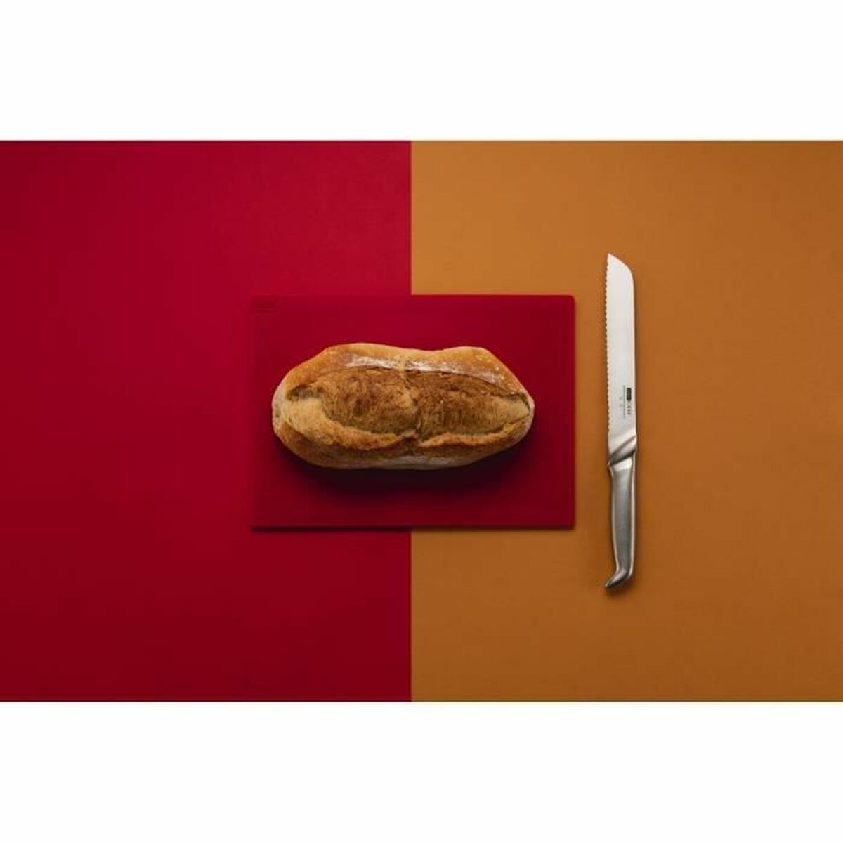 Ein Brotlaib auf einem roten Schneidebrett, ein gezahntes Messer und ein Hintergrund, der zwischen Rot und Orange geteilt ist.