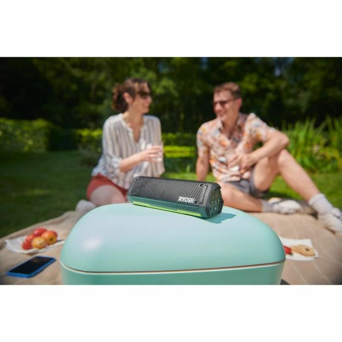 Un altoparlante Ryobi è su un frigorifero. Due persone sono sedute sullo sfondo. Mele e un telefono sono su una coperta da picnic.