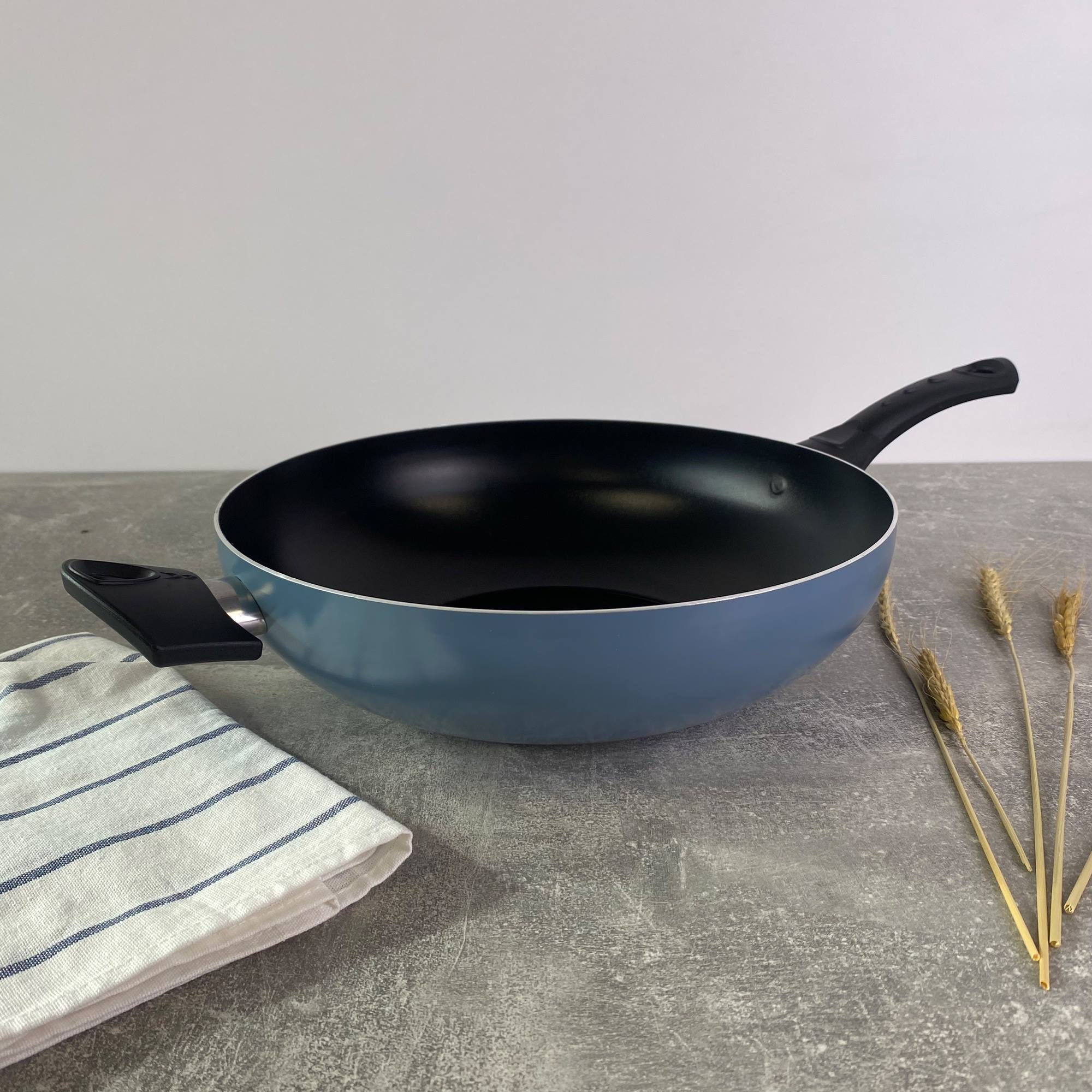 Wok azul sobre una superficie gris con un paño a rayas blanco y azul y trigo. Fondo blanco.