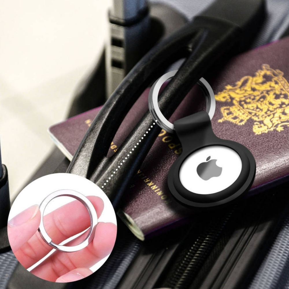 Ein Apple AirTag in einem schwarzen Gehäuse mit einem silbernen Ring, neben einem Reisepass auf einem Koffer. Eine Hand hält einen silbernen Ring.