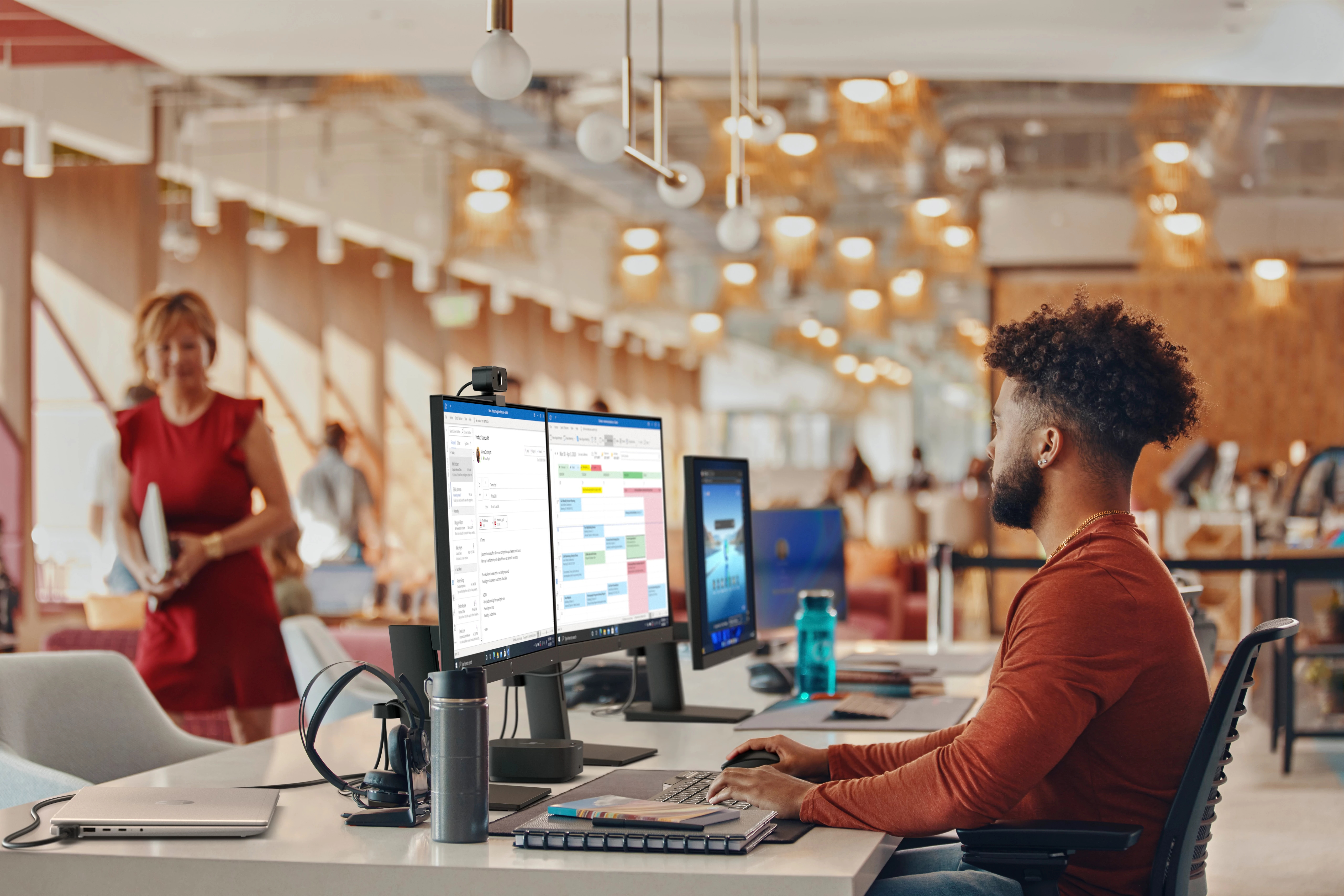 Mężczyzna w biurze, pracujący na komputerze. Kobieta w czerwonej sukience w tle. Wnętrze biura z wieloma ekranami.