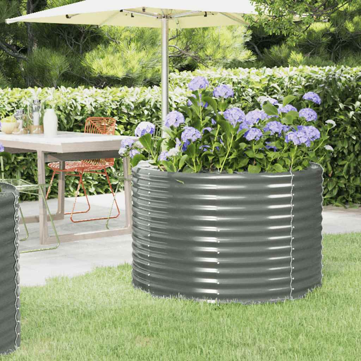 Eine Gartenszene mit einem Metallpflanzer voller lila Hortensien. Im Hintergrund stehen ein Tisch und Stühle unter einem weißen Sonnenschirm.