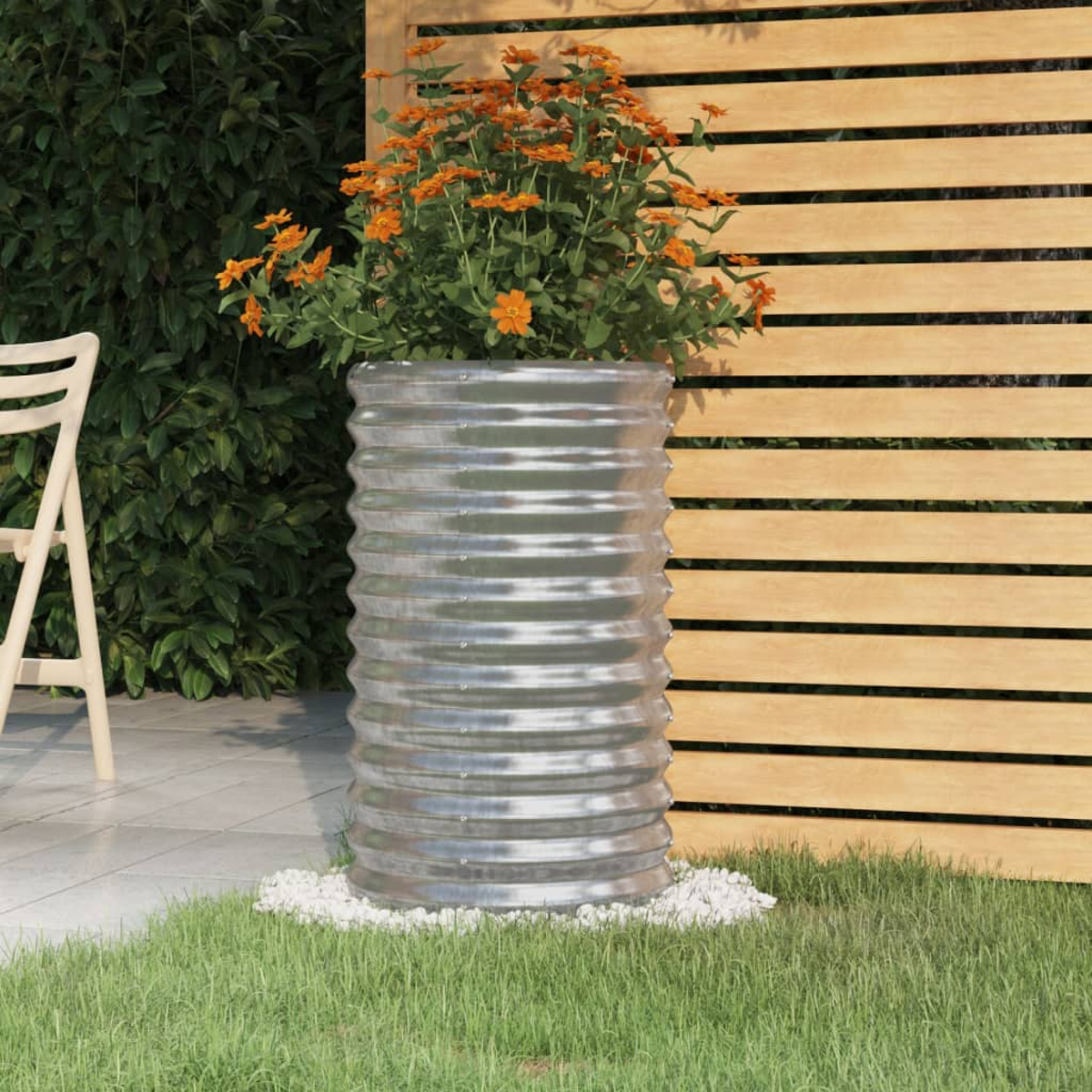 Ein großer, gewellter Metallpflanzer mit orangefarbenen Blumen steht auf einem Bett aus weißen Steinen in einem Garten.