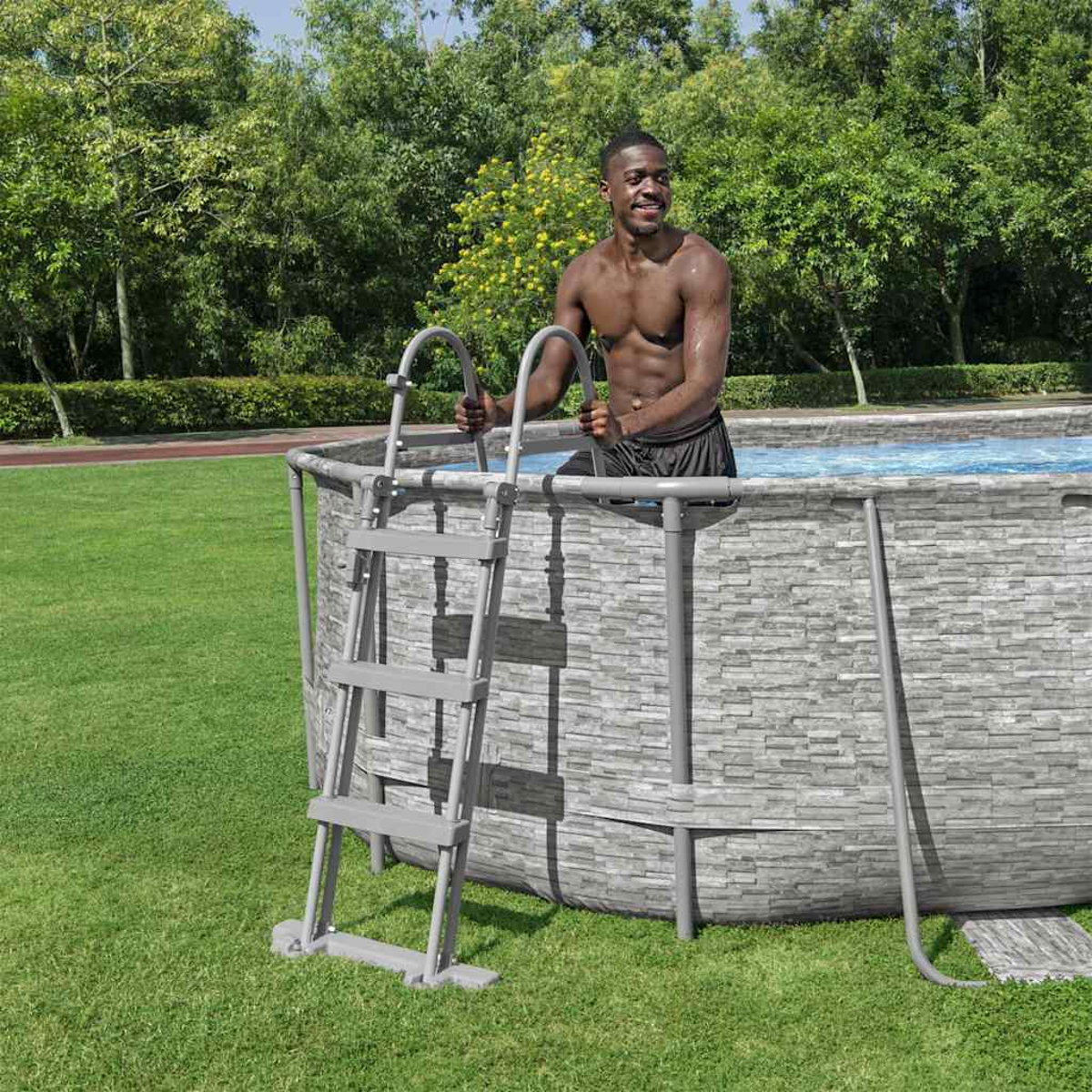 Ein Mann steigt über eine Leiter aus einem Pool. Der Pool ist von einer steinartigen Wand umgeben. Grünes Gras und Bäume bilden den Hintergrund.