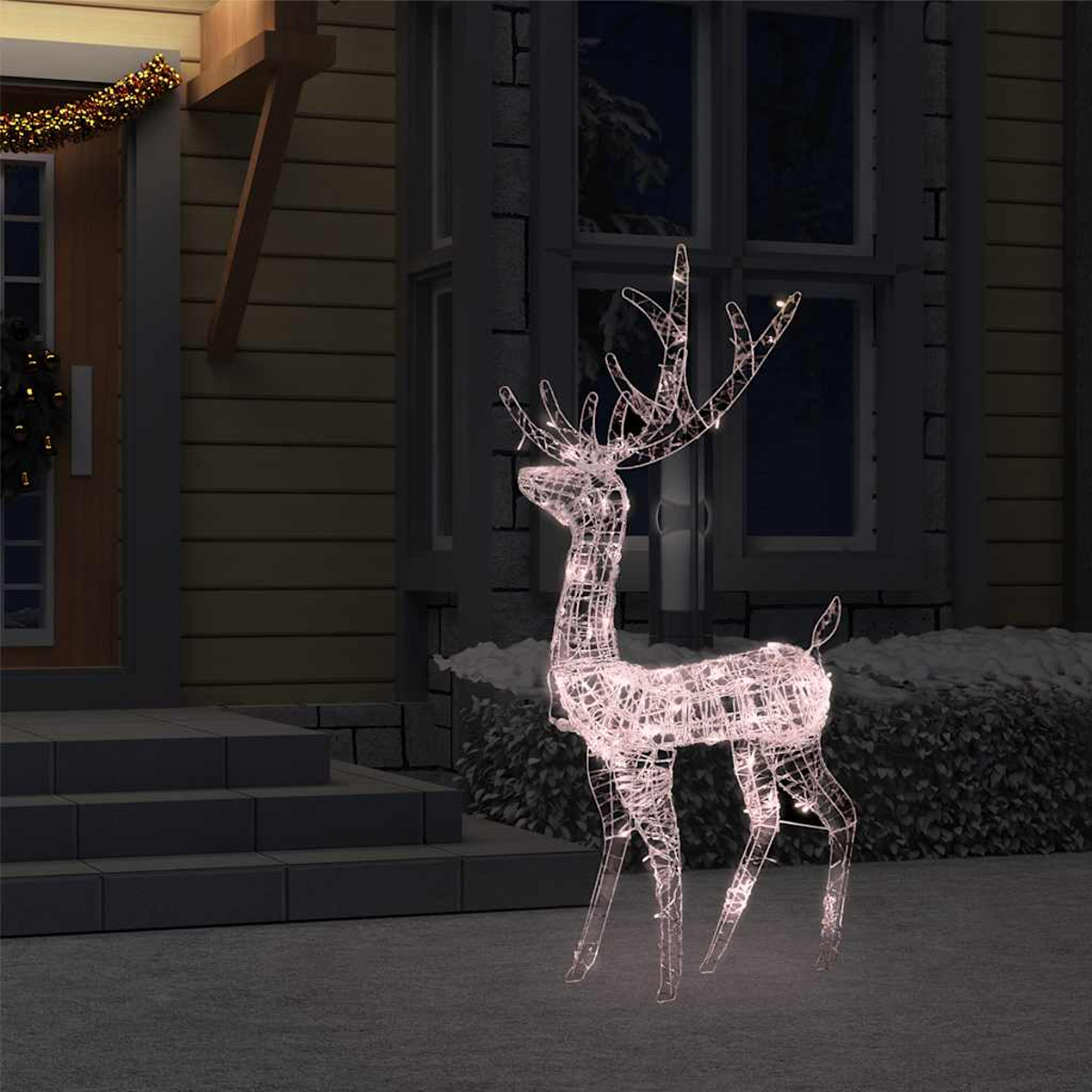Eine beleuchtete Rentierdekoration steht auf einem schneebedeckten Rasen vor einem Haus. Weihnachtsbeleuchtung schmückt das Haus und die Tür.