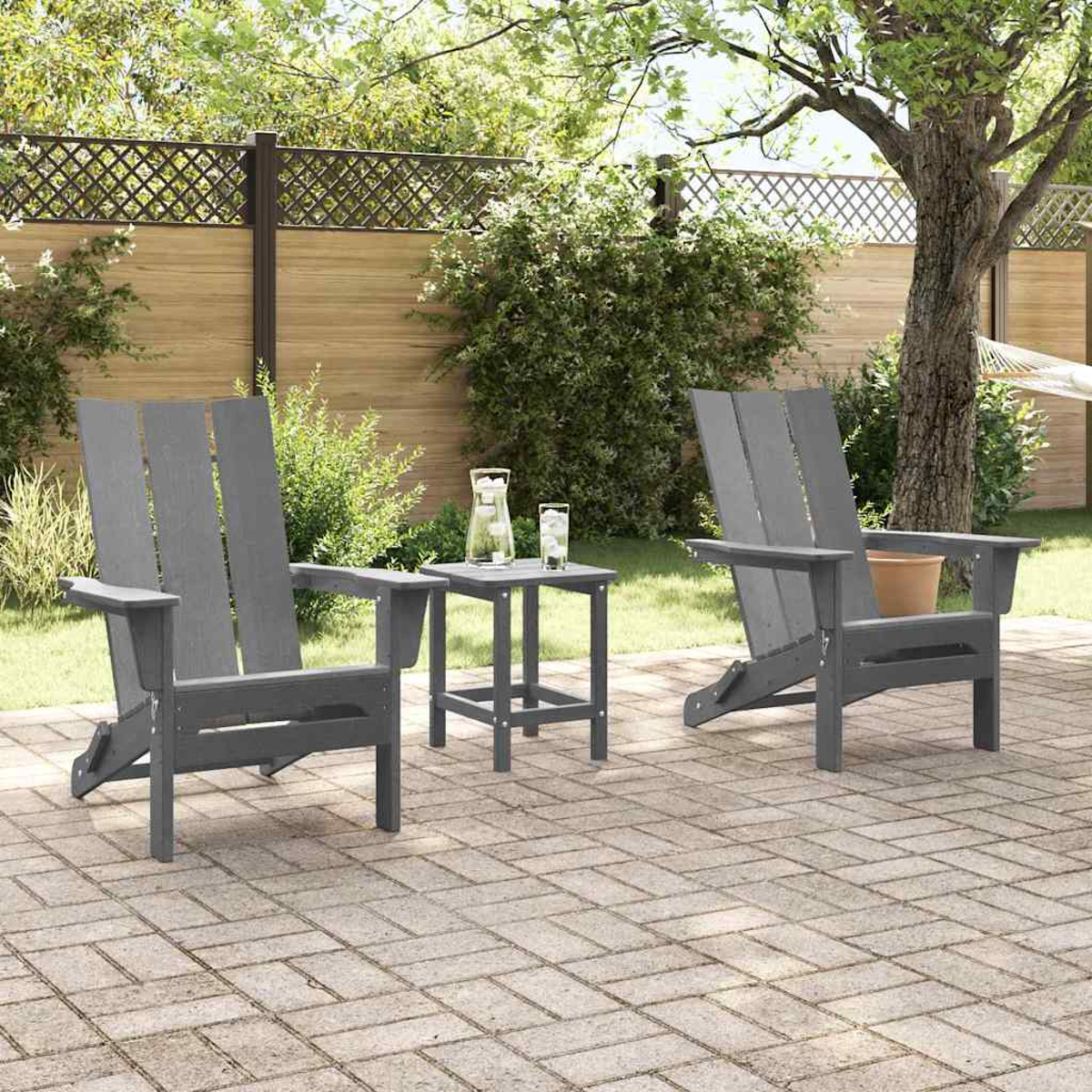 Zwei graue Adirondack-Stühle mit einem kleinen Tisch auf einer Ziegelterrasse. Ein Krug und Gläser stehen auf dem Tisch. Grün im Hintergrund.