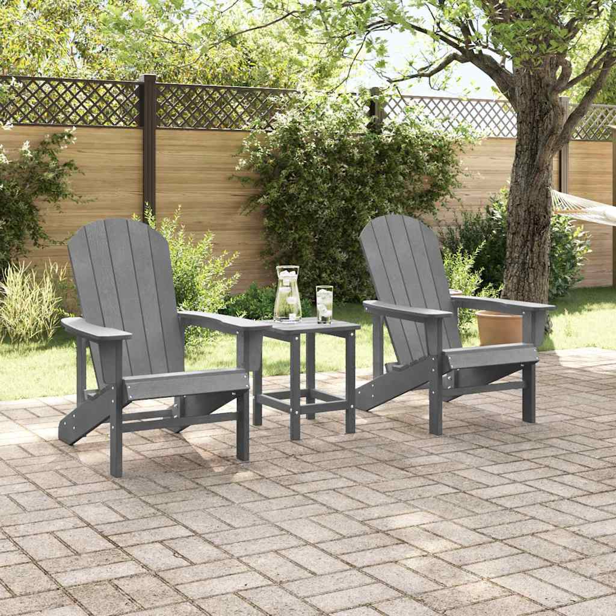 Zwei graue Adirondack-Stühle mit einem kleinen Tisch, Getränken auf einer Ziegelterrasse. Grünes Gras und Bäume im Hintergrund.