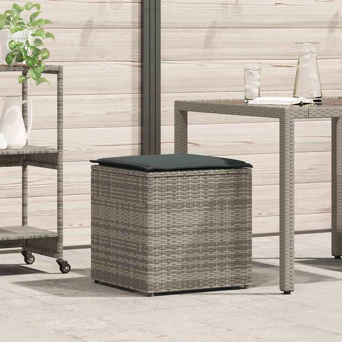 Ein gewebter, grau-quadratischer Hocker mit schwarzem Kissen. Ein Tisch und ein Wagen mit Pflanzen im Hintergrund.
