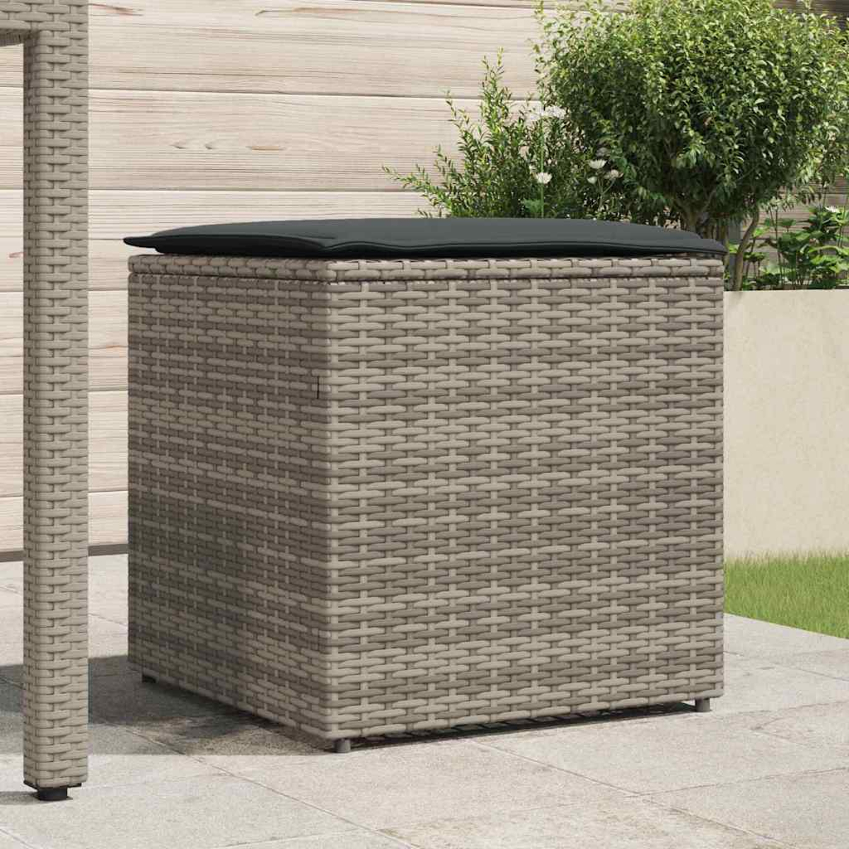 Eine graue Weidenkorb-Aufbewahrungsbox mit einem dunklen Kissen oben, auf einer Terrasse. Ein Tischbein links, Grünzeug im Hintergrund.