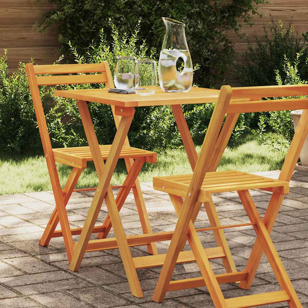 Holztisch im Freien mit zwei Stühlen, einem Wasserkrug und zwei Gläsern. Die Szene spielt sich in einem Garten mit grünem Laub ab.