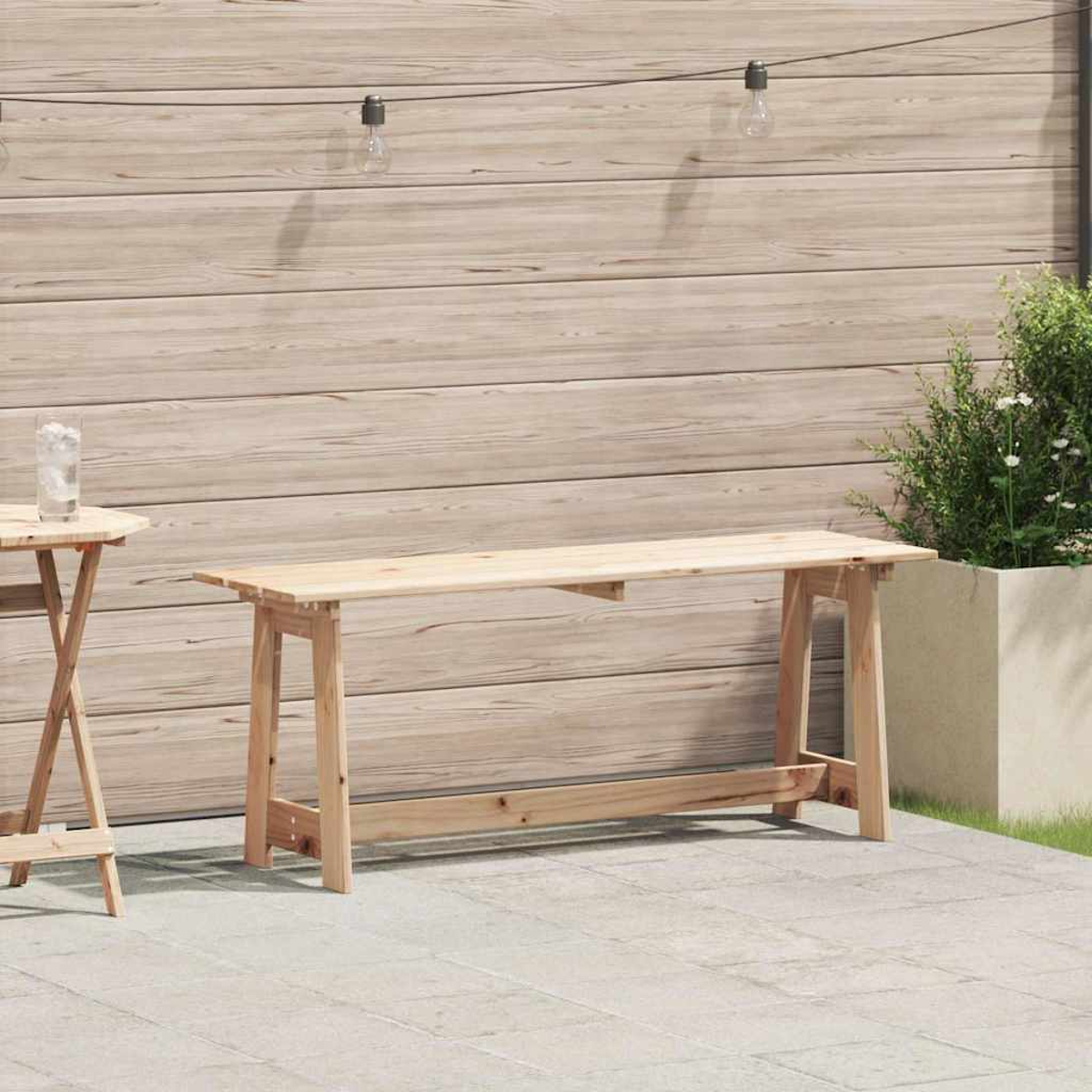 Eine Holzbank und ein kleiner Tisch stehen auf einer Terrasse vor einer Holzwand. Ein Pflanzgefäß mit Grün ist ebenfalls vorhanden.