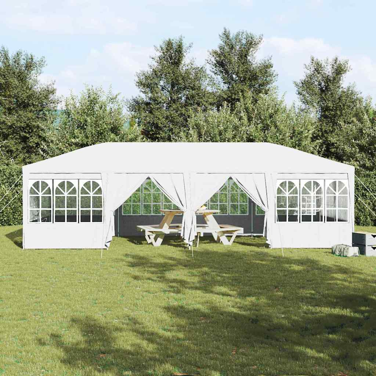 Una carpa blanca está instalada en un césped verde, con mesas de picnic en el interior. Los árboles forman el fondo y el cielo es visible.