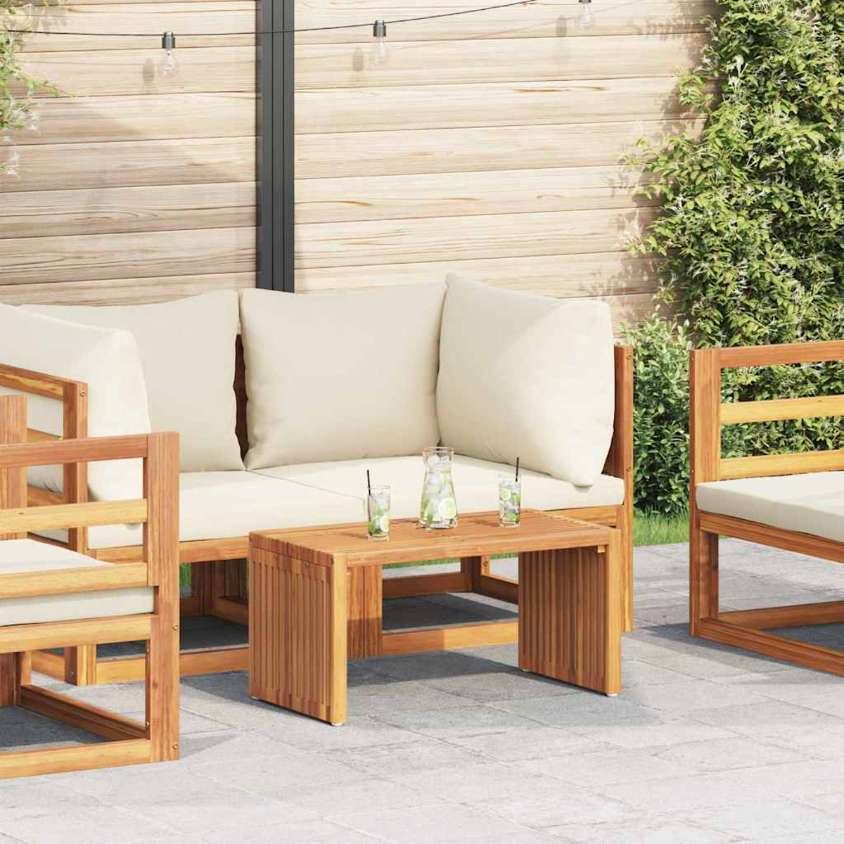 Holz-Sitzbereich im Freien. Cremefarbene Kissen auf einem Sofa, zwei Stühlen und einem Tisch mit Getränken. Holz-Hintergrund und Grün.