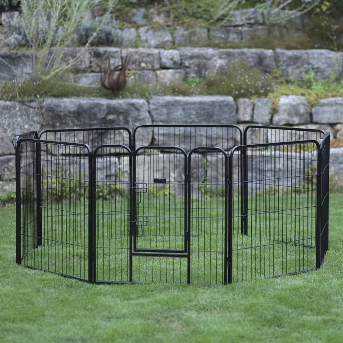 Ein schwarzer Metall-Haustierauslauf auf grünem Gras, mit einer Steinmauer und Grünzeug im Hintergrund.