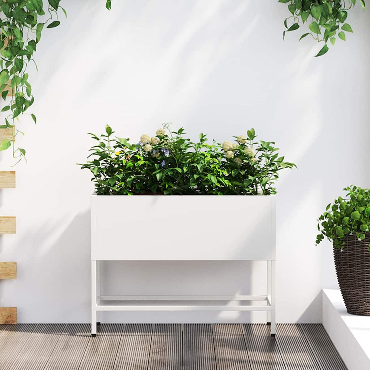 Una fioriera bianca con piante verdi e fiori è su una terrazza in legno. L'edera pende dall'alto. Un'altra pianta in vaso è a destra.