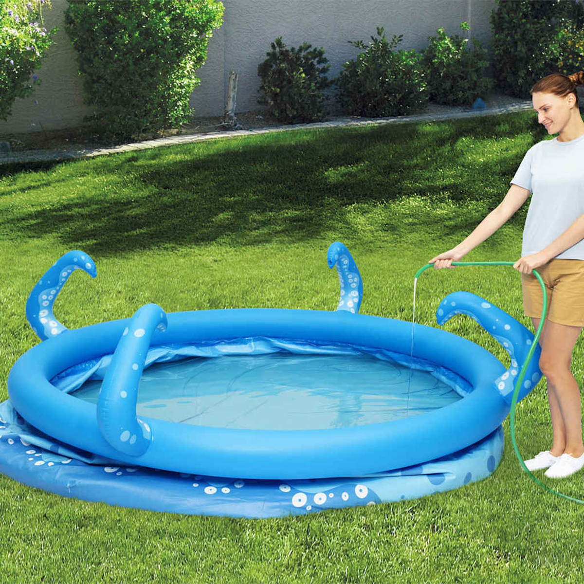 Eine Frau füllt einen blauen, oktopusförmigen aufblasbaren Pool mit Wasser aus einem grünen Schlauch auf Rasen.