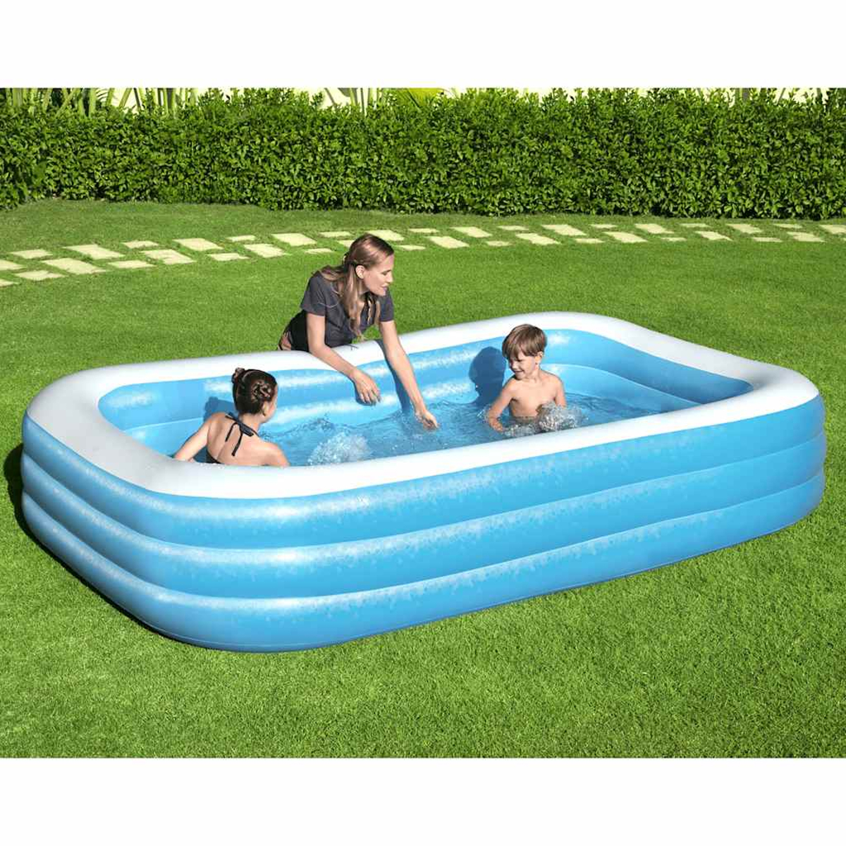 Eine Frau und zwei Kinder befinden sich in einem aufblasbaren blauen Pool auf grünem Gras. Im Hintergrund befindet sich eine Hecke.