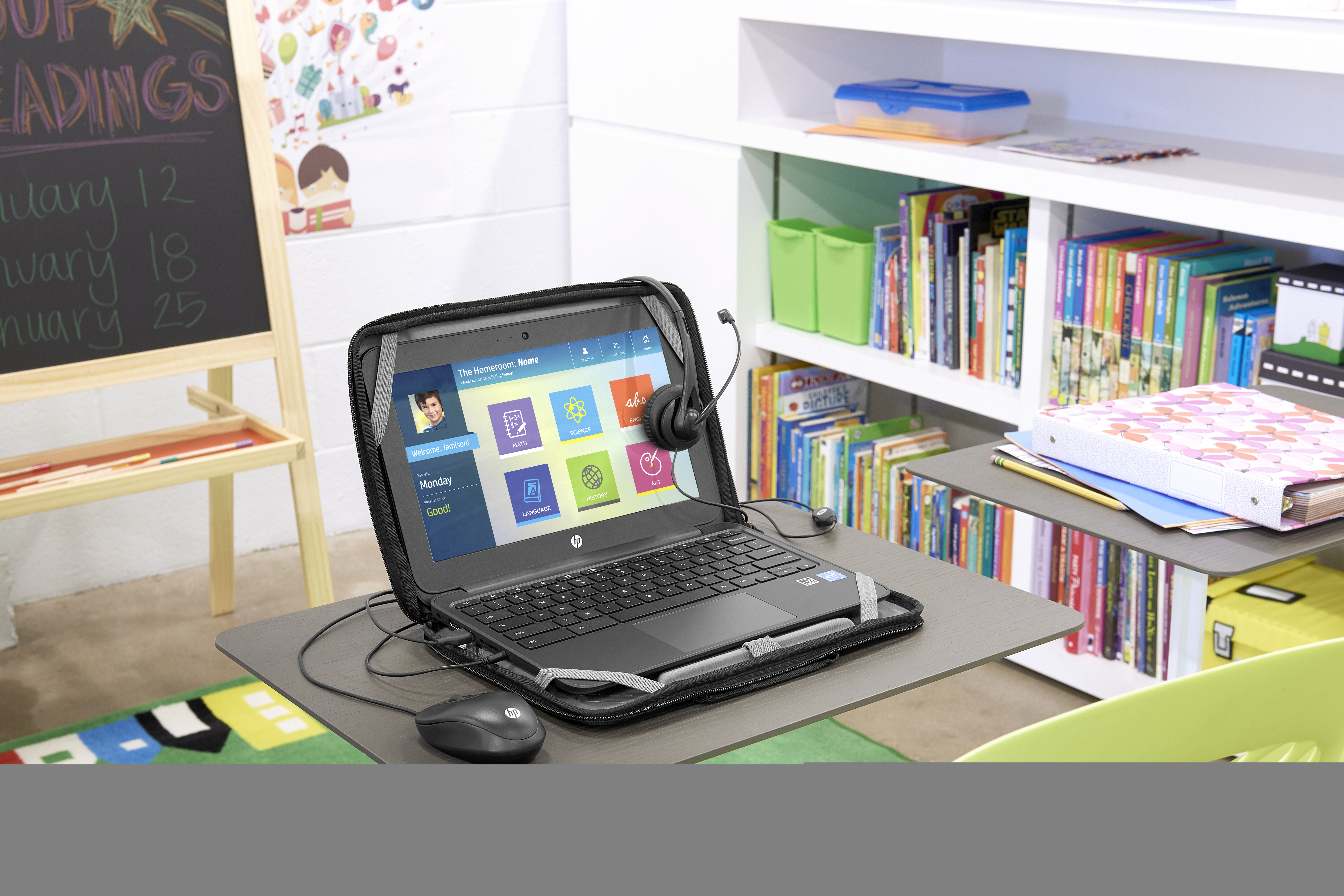 Laptop su una scrivania con mouse e cuffie. Una lavagna dietro e una libreria sullo sfondo.