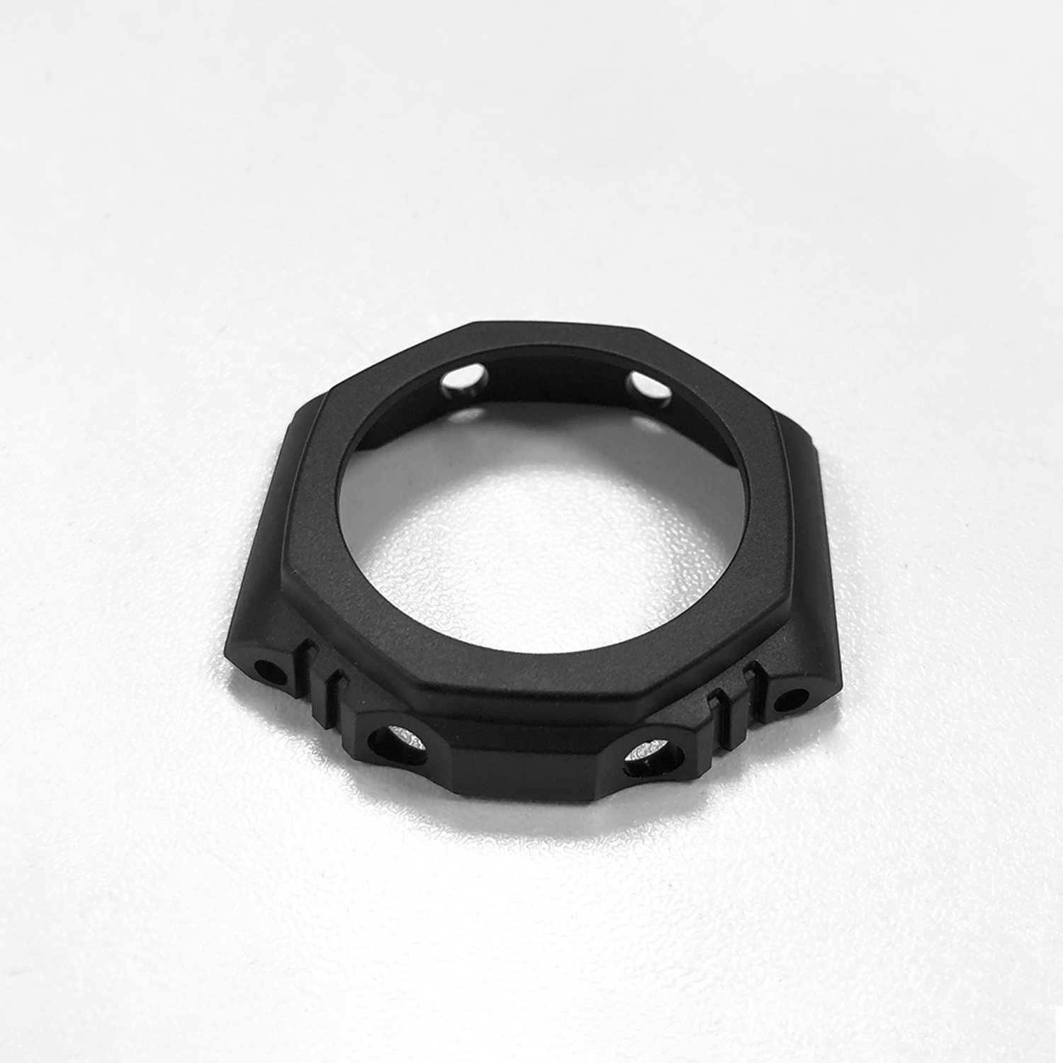 Eine schwarze Uhrenlünette liegt auf einer weißen Oberfläche. Sie hat eine achteckige Form mit Löchern und besteht aus mattem Material.
