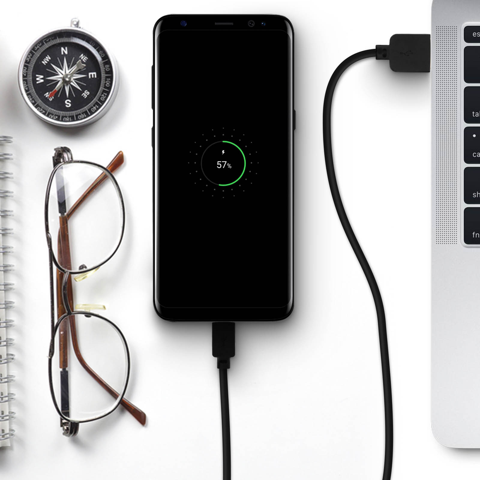 Un telefono si sta caricando con un cavo USB. Una bussola, occhiali e un laptop sono anche sulla superficie bianca.
