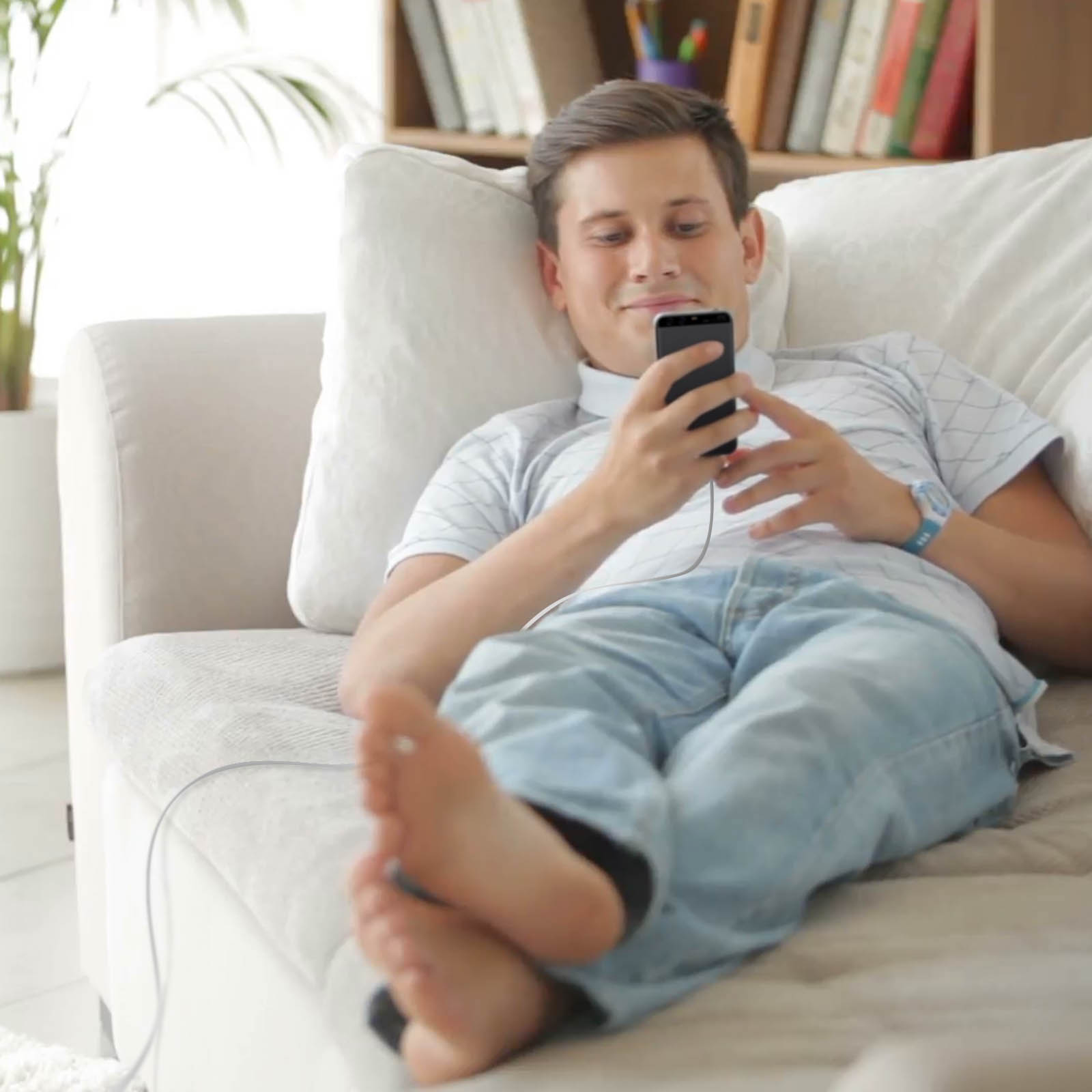 Un uomo è sdraiato su un divano, usa un telefono. Indossa jeans e camicia chiara. Piedi nudi in primo piano.