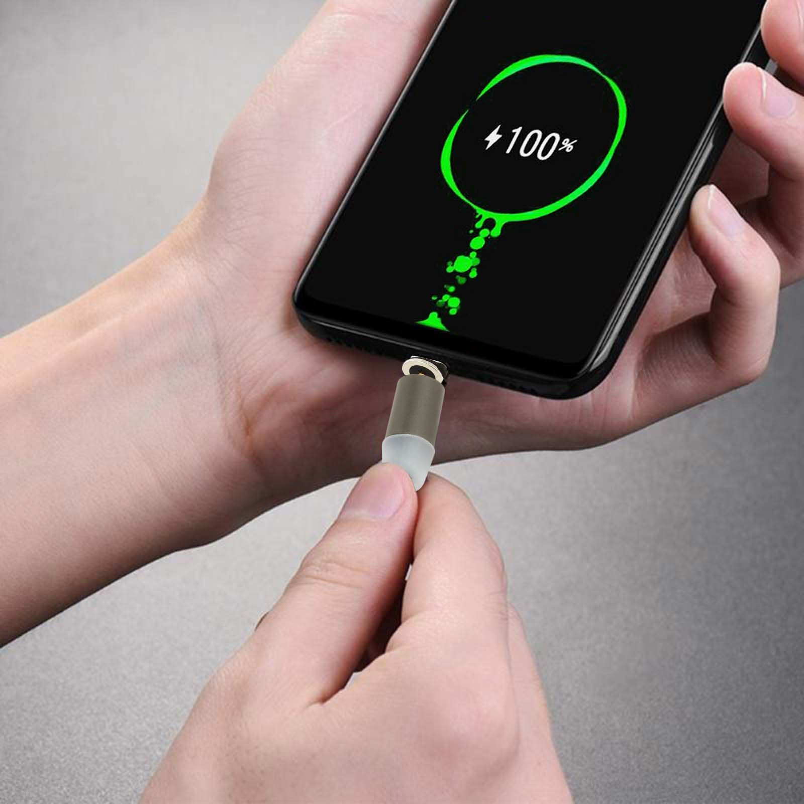 Un telefono mostra la carica al 100%. Una mano tiene un caricabatterie collegato al telefono.
