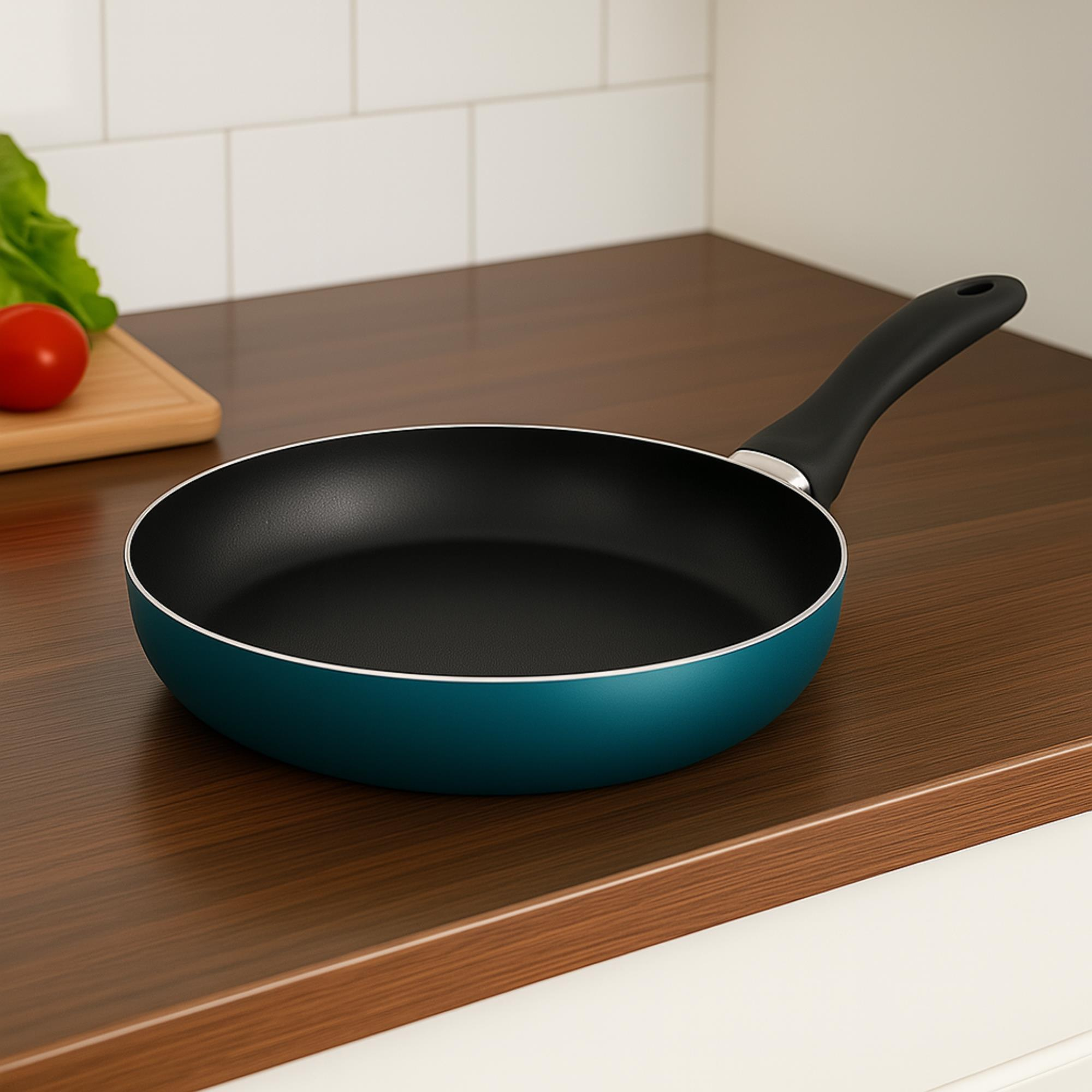 Una sartén azul con mango negro sobre encimera de madera. Tomate y lechuga sobre tabla de cortar.