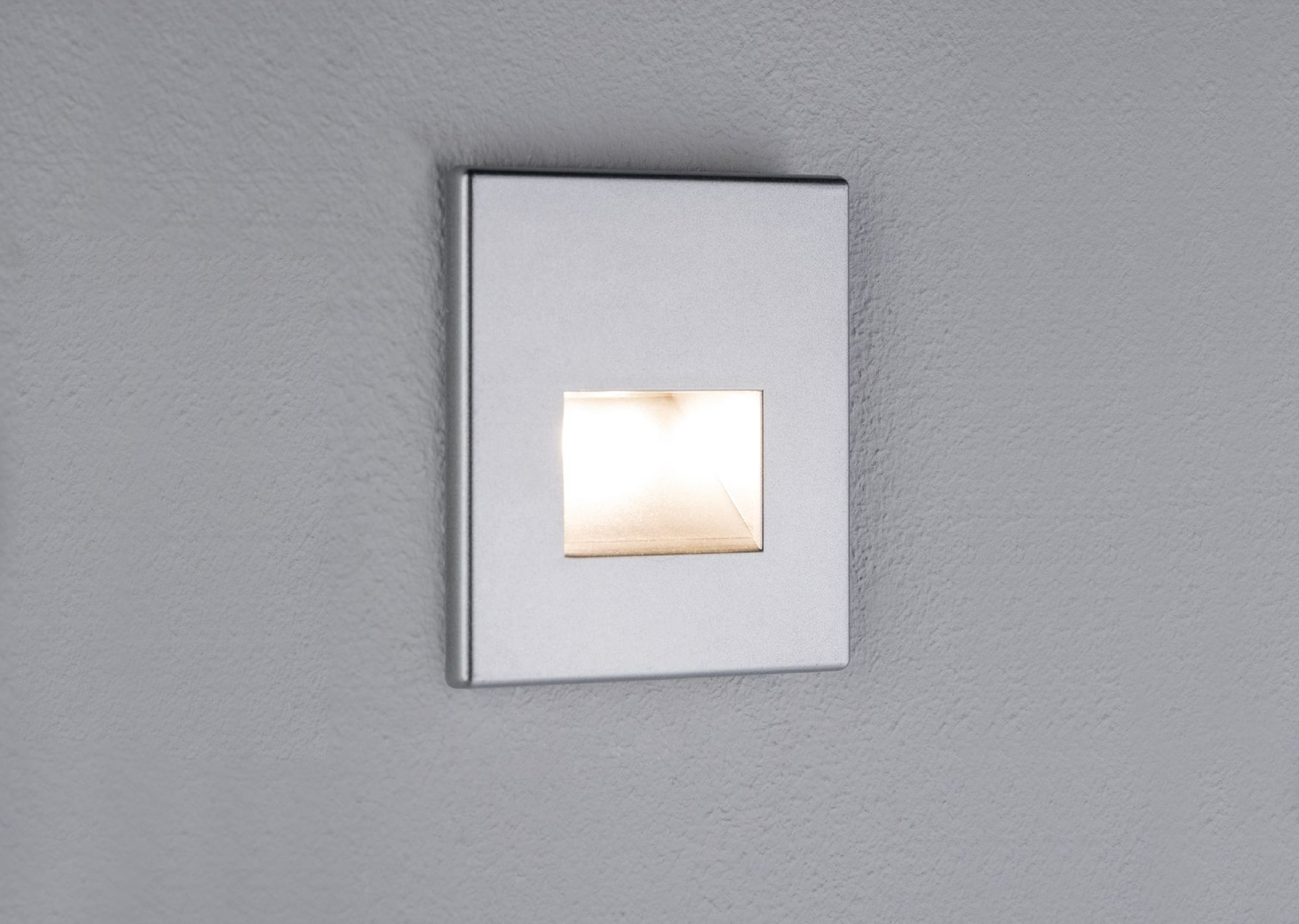 Un luminaire carré argenté est monté sur un mur gris texturé. La lumière à l'intérieur brille.