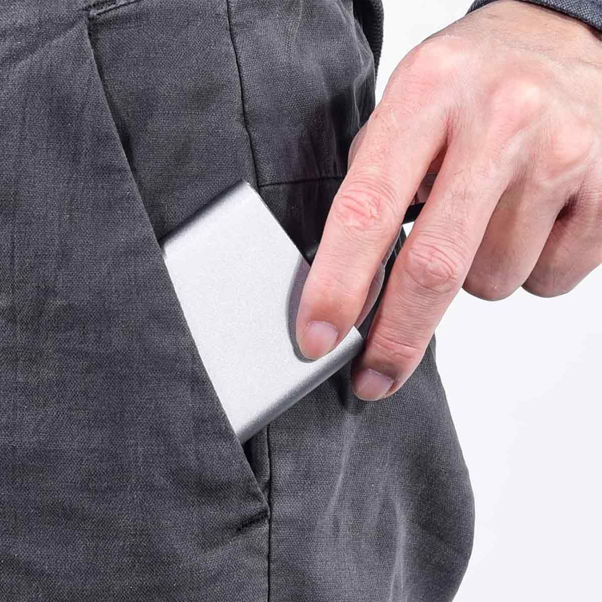 Eine Hand greift in eine Tasche und umfasst ein silbernes, rechteckiges Gerät. Die Tasche gehört zu einer grauen Hose.