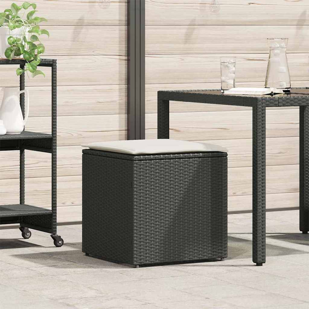Dunkelgraue, gewebte Aufbewahrungshocker mit weißem Kissen auf der Terrasse. Tisch, Wagen und Holzwand im Hintergrund.