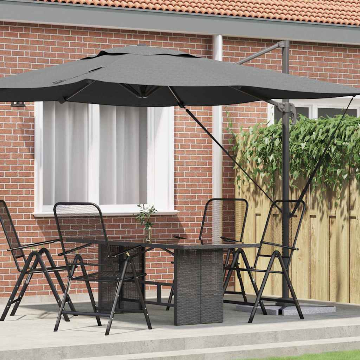 Set da patio con ombrellone. Tavolo e sedie sono neri. Muro di mattoni rossi e recinzione in legno sullo sfondo. Ombrellone grigio ombreggia il set.