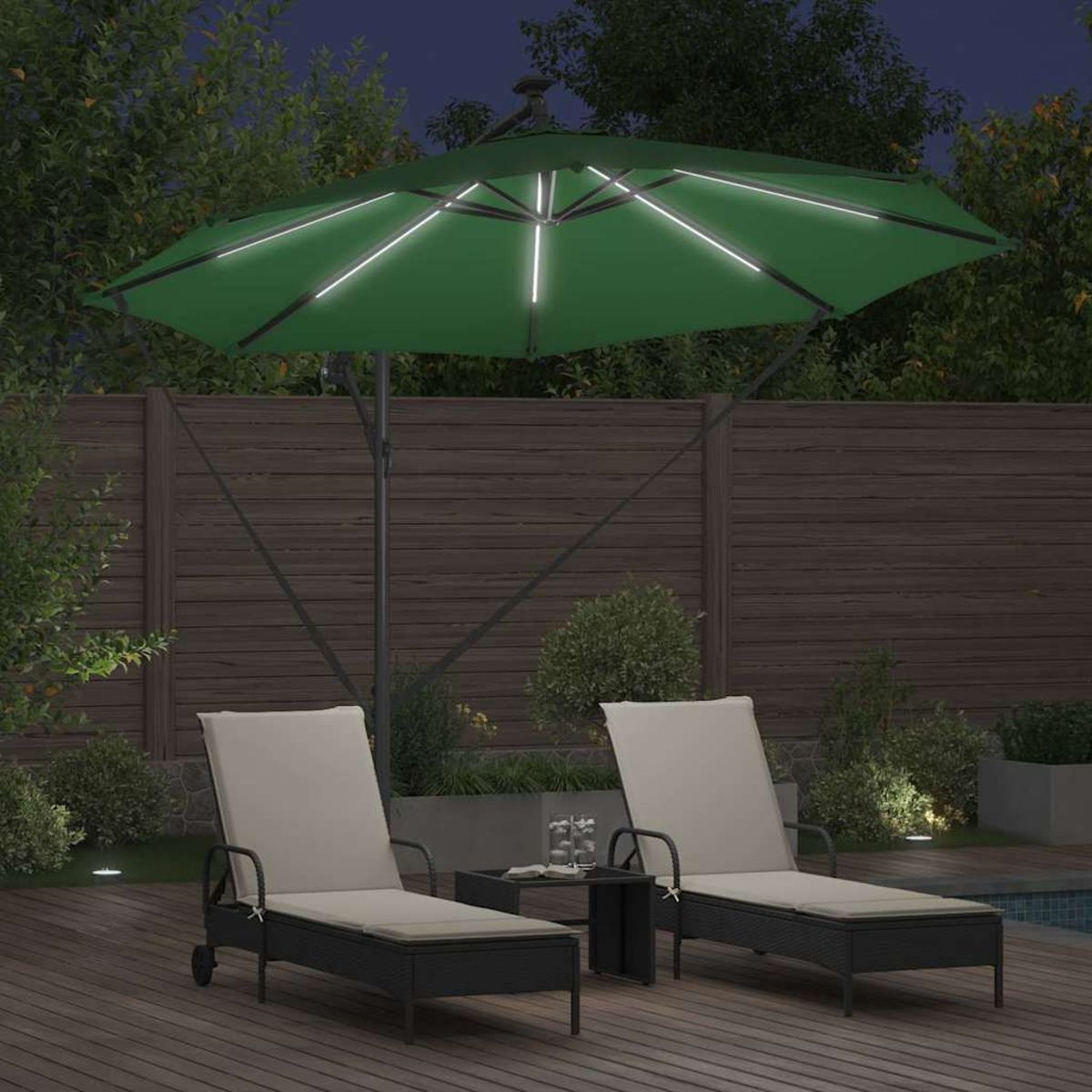 Scena notturna: Ombrello verde illuminato sopra due sedie a sdraio. Un tavolino con bicchieri tra loro su una terrazza di legno.