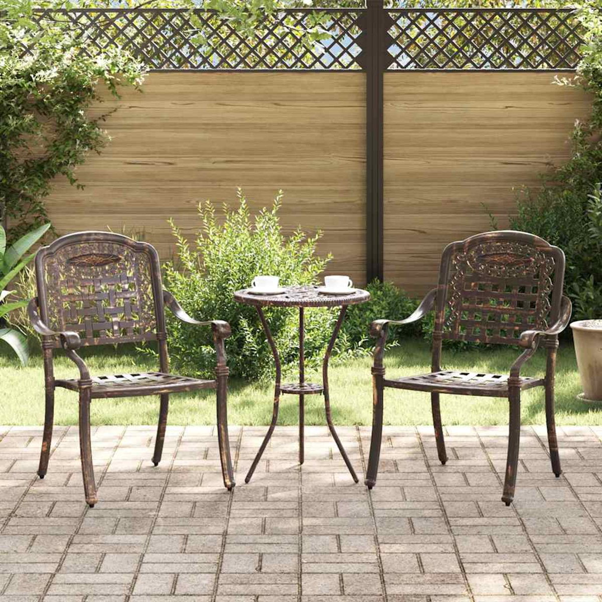 Zwei verzierte Metallstühle und ein kleiner Tisch auf einer Terrasse. Im Hintergrund ein Holzzaun. Zwei Tassen stehen auf dem Tisch.