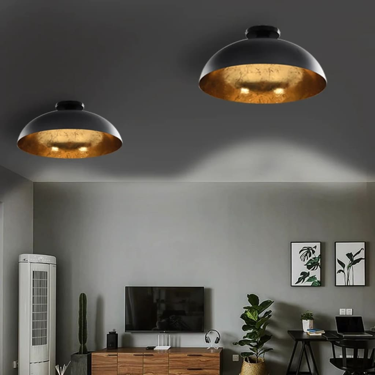 Zwei schwarze und goldene Pendelleuchten hängen an einer grauen Decke. Darunter ist ein Wohnzimmer mit Pflanzen, einem Fernseher und einem Schreibtisch zu sehen.