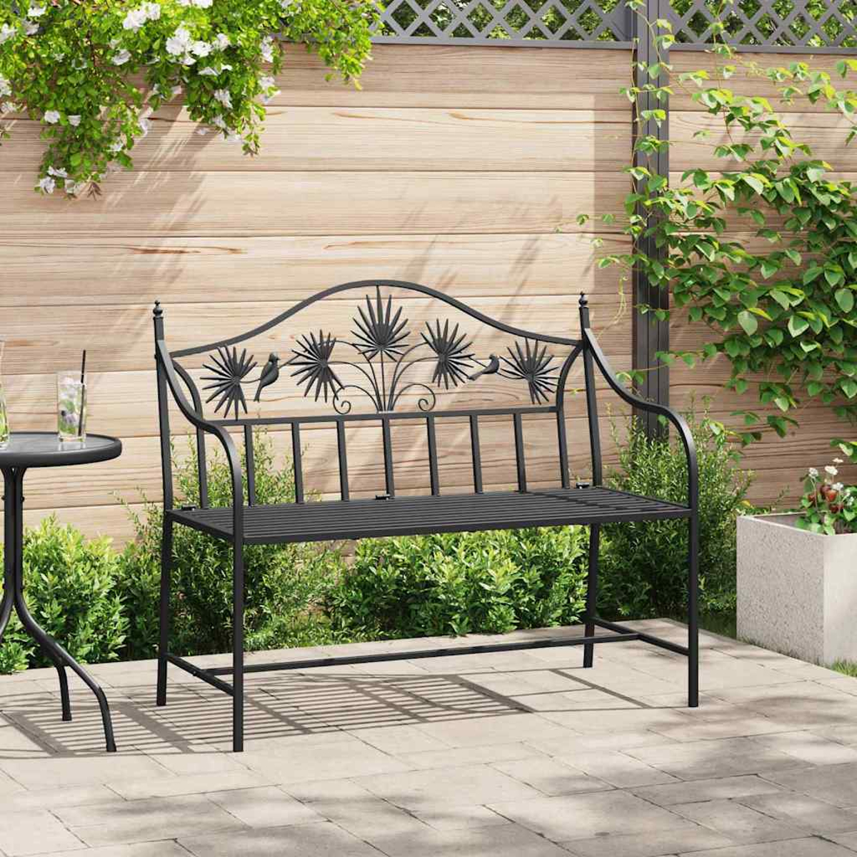 Schwarze Metallbank mit dekorativer Rückenlehne in einem Garten. Kleiner Tisch mit Getränk links und Grünzeug im Hintergrund.