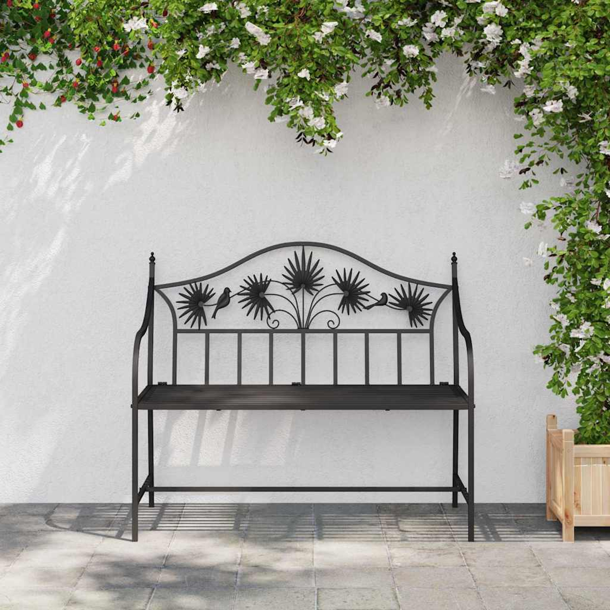 Eine schwarze Metallbank mit Blumen- und Vogelmotiven steht vor einer weißen Wand mit hängenden Blumen. Rechts ist ein Holzpflanzer.