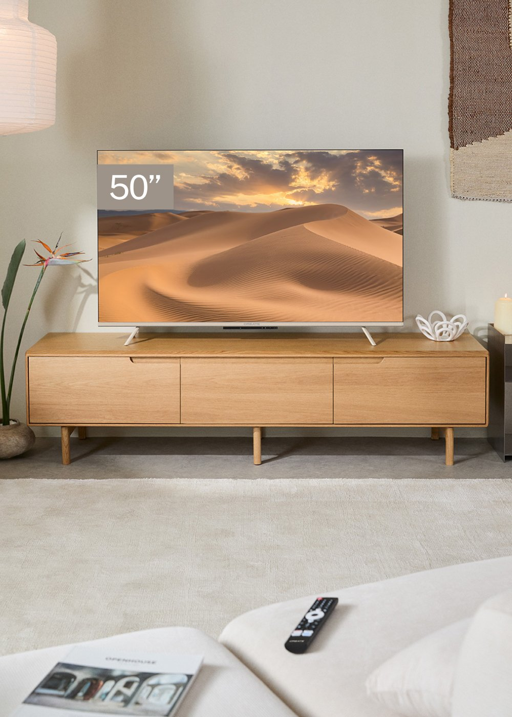 Un televisor de 50 pulgadas muestra un paisaje desértico. Debajo, un mueble de madera sobre una alfombra clara, con mando y libro.