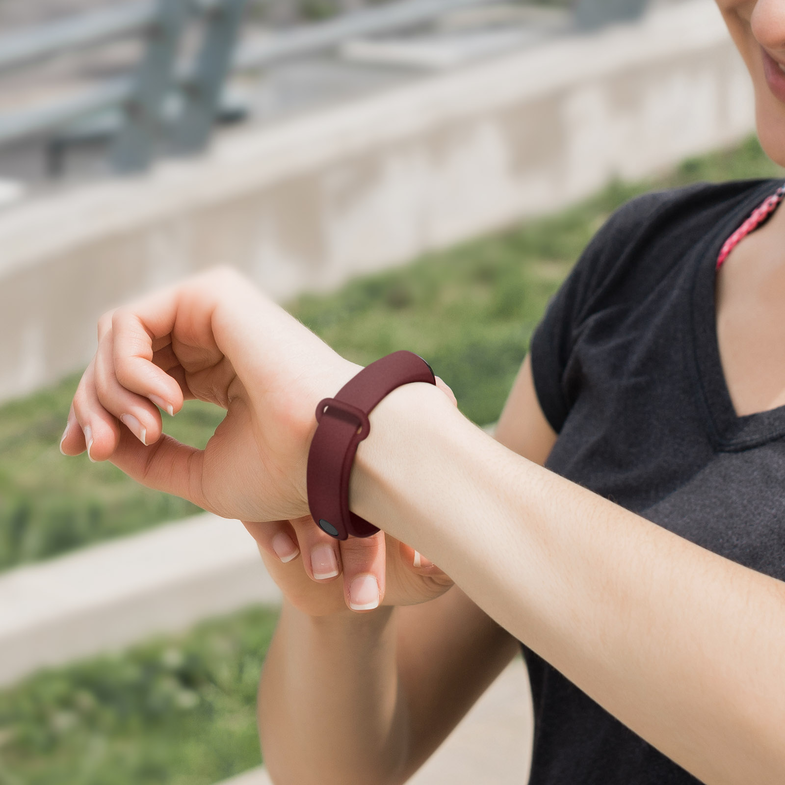 Una persona con un fitness tracker bordeaux al polso, che lo guarda. Lo sfondo è sfocato con verde.