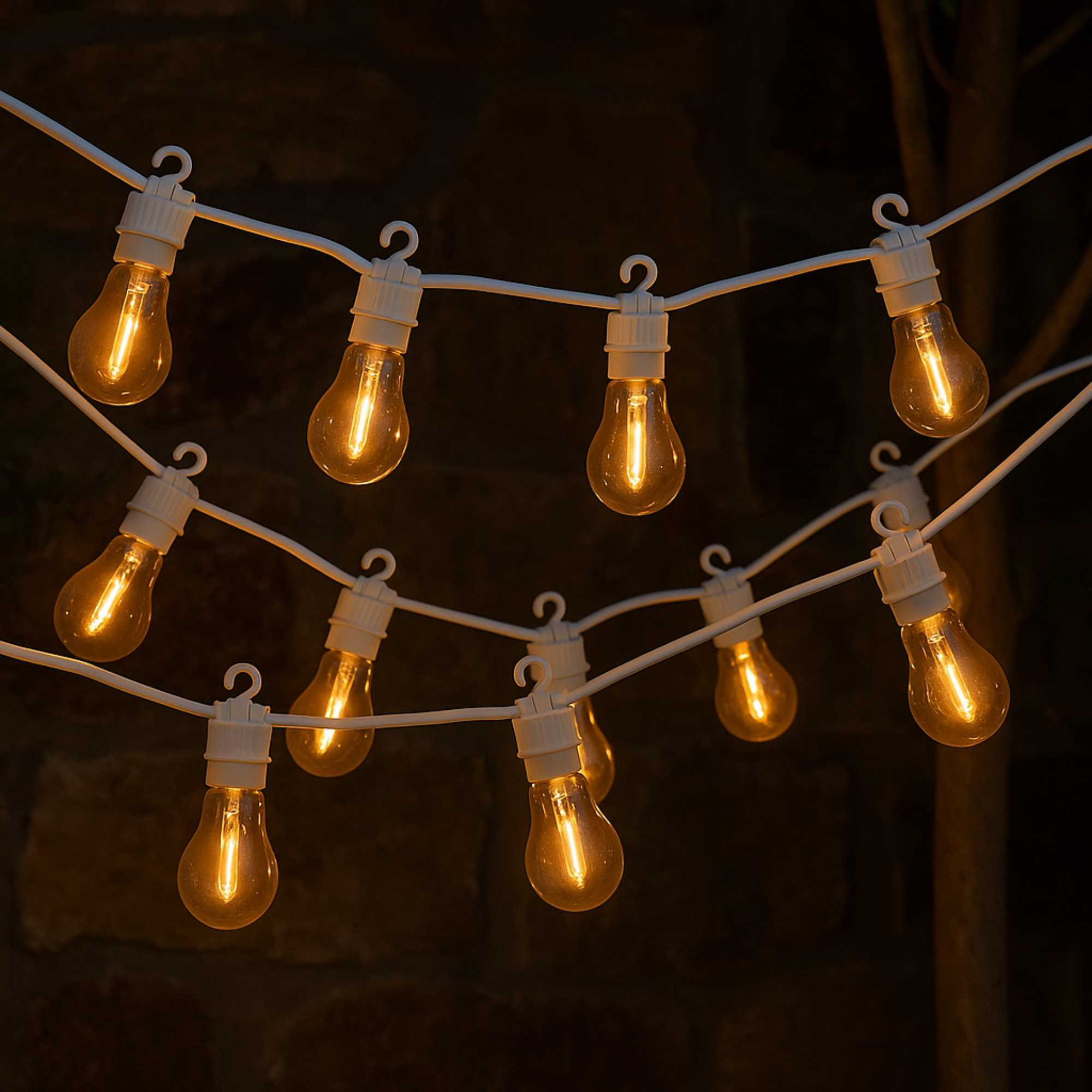 Luces de cadena con bombillas cálidas y brillantes colgando sobre un fondo oscuro. Se ven el cable blanco y los portalámparas.