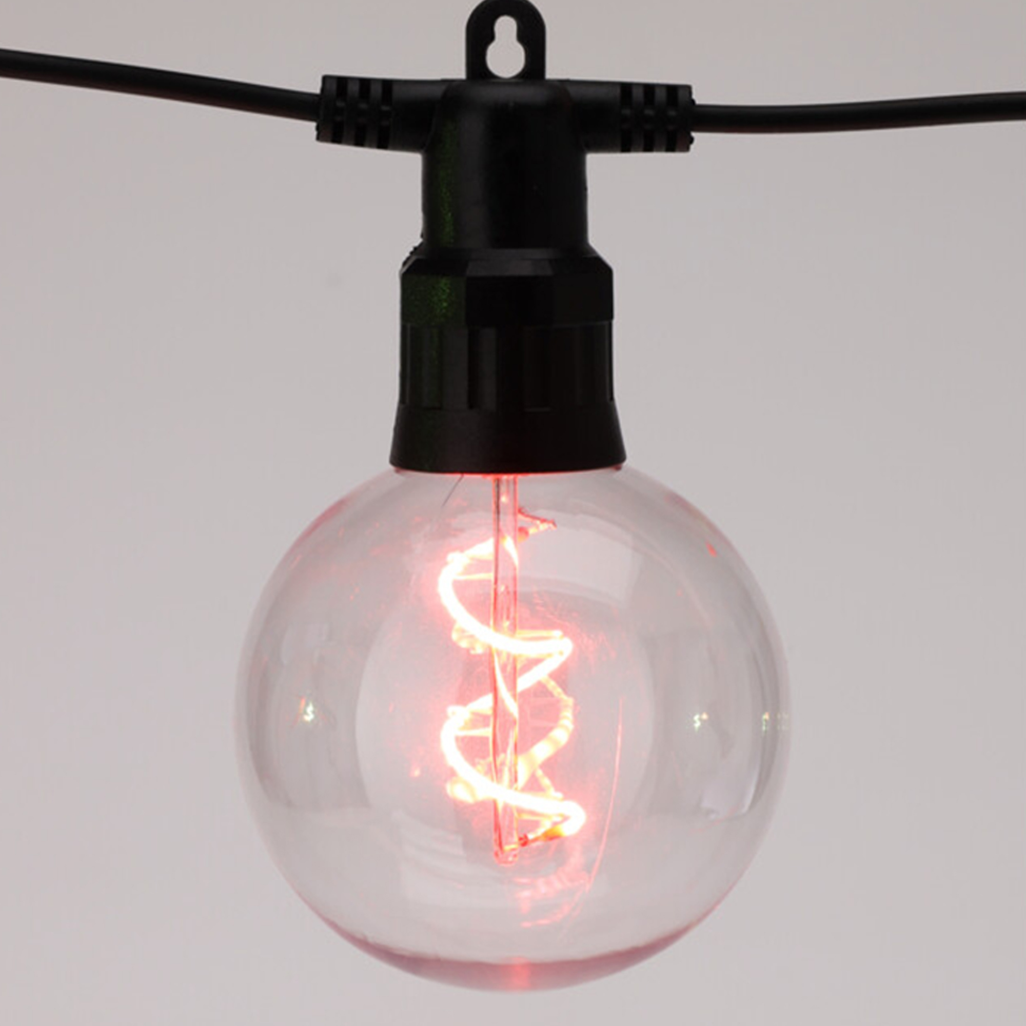 Una bombilla transparente y redonda con un filamento rojo-anaranjado encendido cuelga de un cable negro, sobre un fondo blanco.
