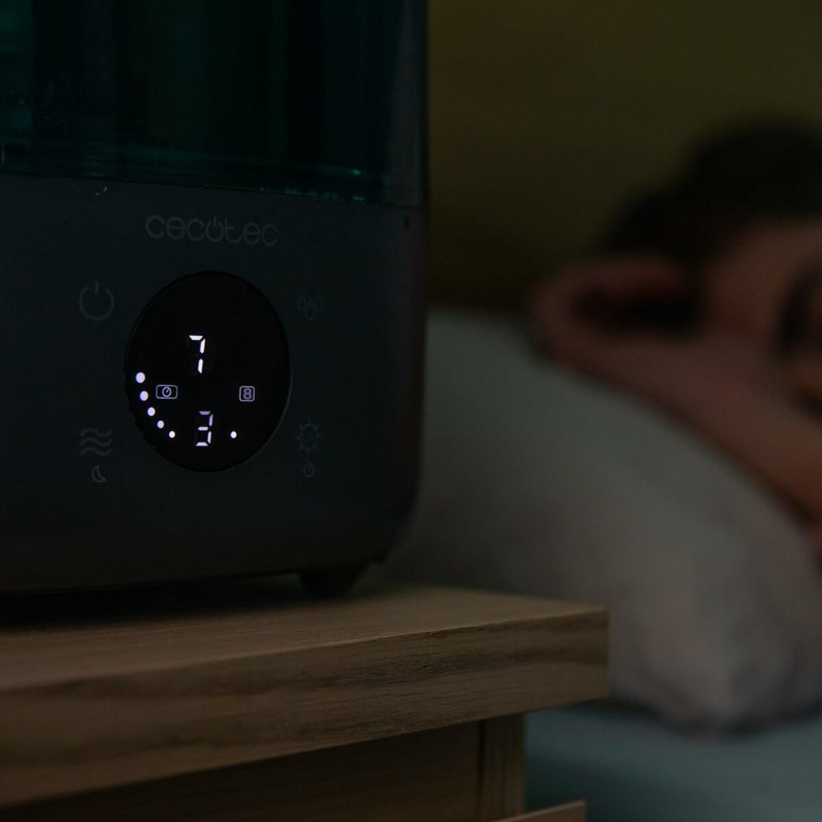Un umidificatore su un comodino di legno mostra l'ora 7:03. Una persona sfocata sullo sfondo sembra dormire.