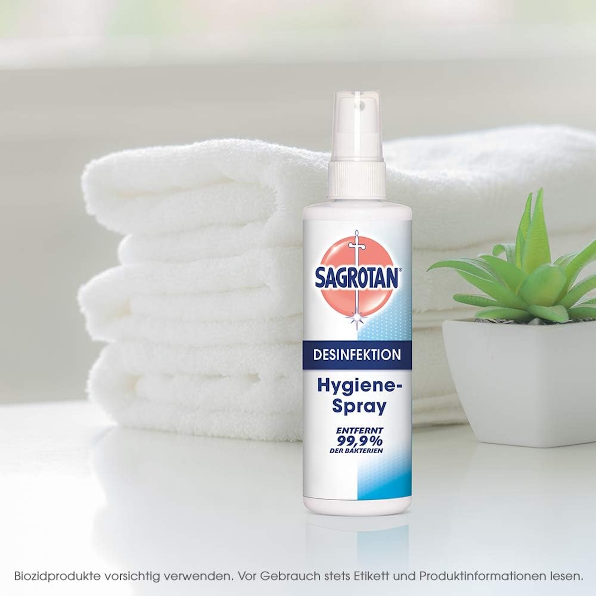 Eine Sagrotan Hygiene-Spray-Flasche steht im Mittelpunkt. Dahinter sind ein Stapel weißer Handtücher und eine kleine Pflanze zu sehen.