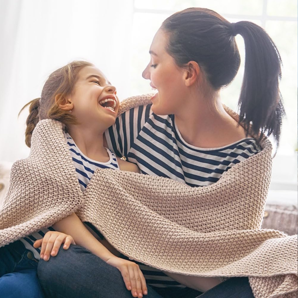 Mutter und Tochter lachen zusammen, in eine beige Decke gehüllt. Beide tragen gestreifte Hemden und sitzen in der Nähe eines Fensters.