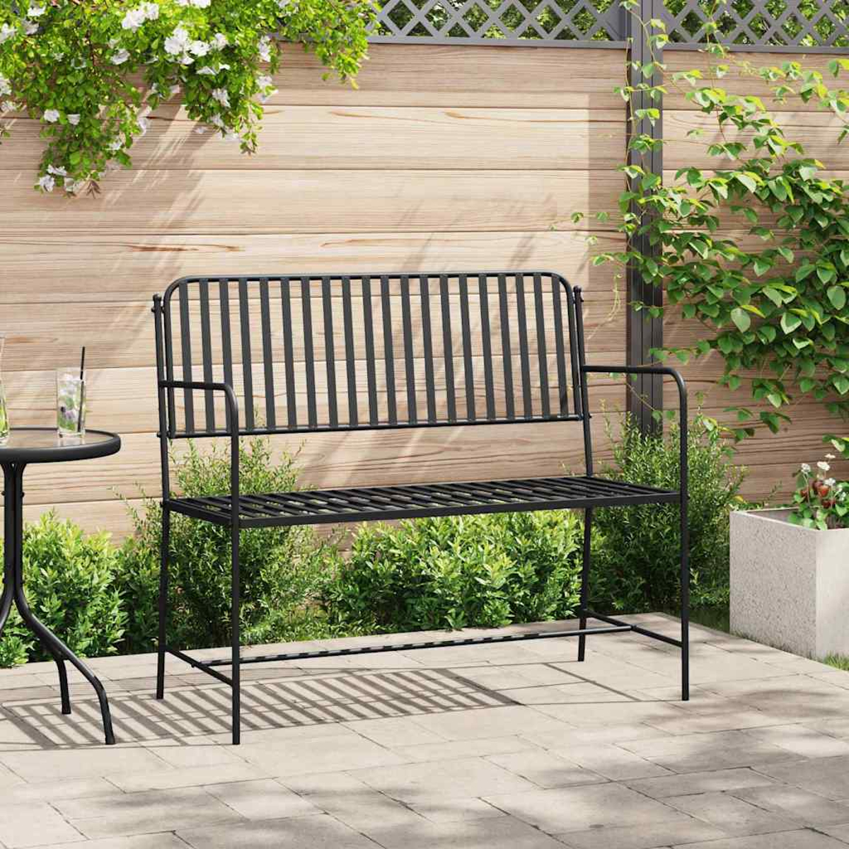 Eine schwarze Metallbank steht auf einer Terrasse. Dahinter befindet sich ein Holzzaun mit Grün und Blumen an den Seiten.