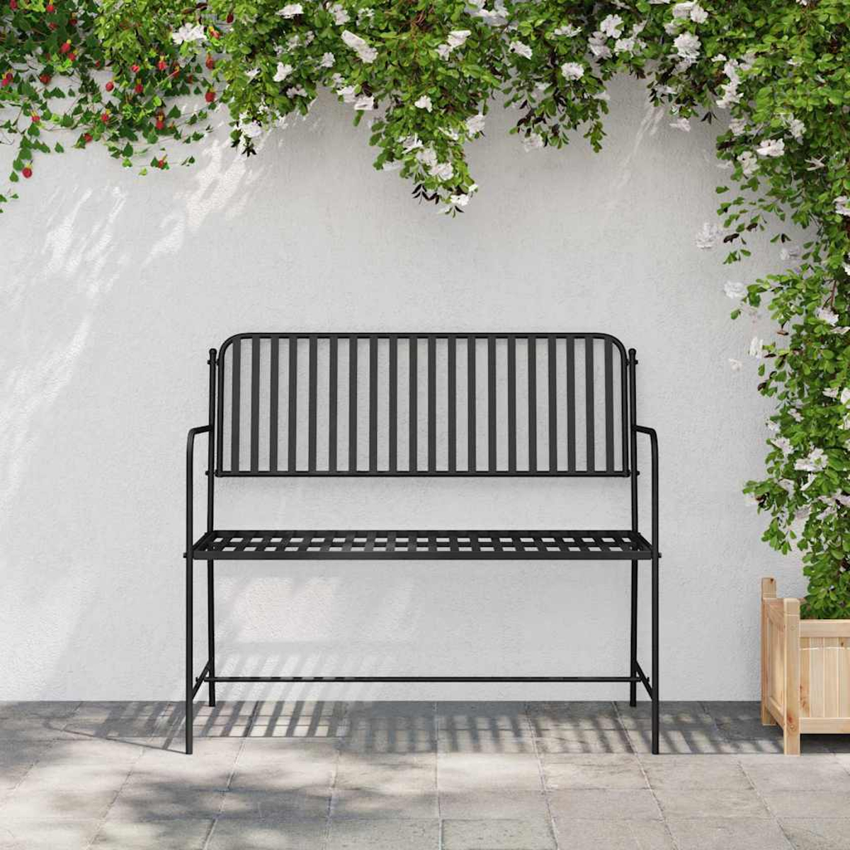 Schwarze Metallbank vor weißer Wand, verziert mit Kletterpflanzen und Blumen, auf einer gepflasterten Terrasse.