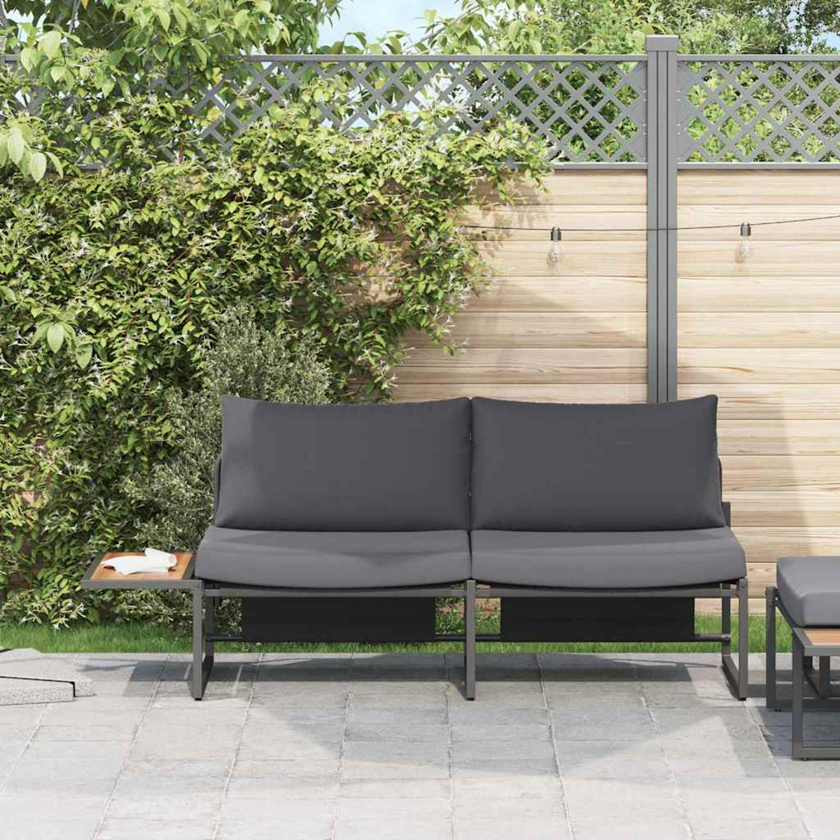 Szene im Freien. Graues Sofa mit zwei Kissen. Holzzaun mit Kletterpflanzen. Beistelltisch mit einer Tissue-Box. Gepflasterte Terrasse.