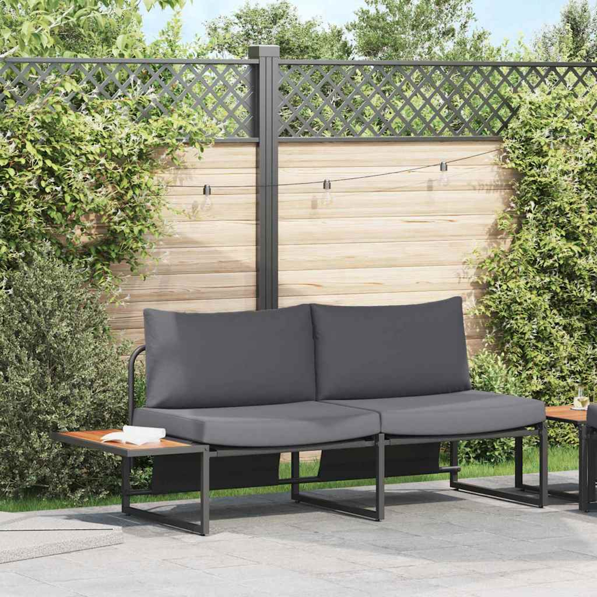 Graues Outdoor-Sofa mit Holztischen. Ein Holzzaun mit Kletterpflanzen bildet den Hintergrund.
