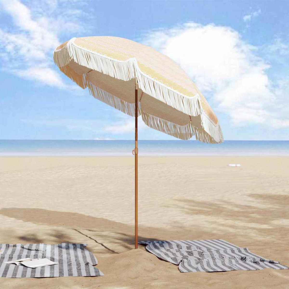 Scena sulla spiaggia con ombrellone a righe, due coperte sulla sabbia e l'oceano sullo sfondo. Il cielo è blu con nuvole.