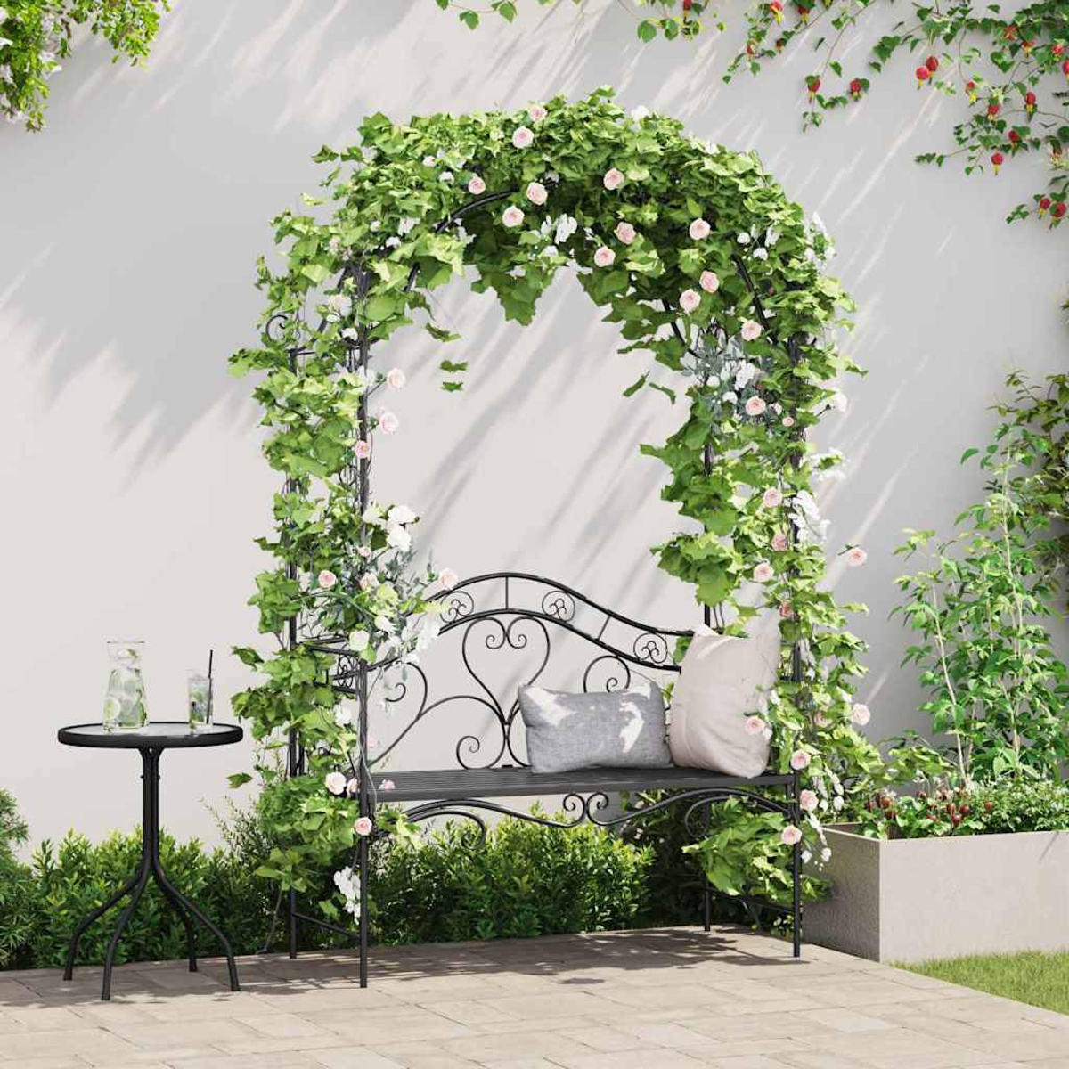 Una panchina in ferro battuto con cuscini sotto un arco floreale. Un tavolino con bevande è a sinistra. Verde e fiori circondano.